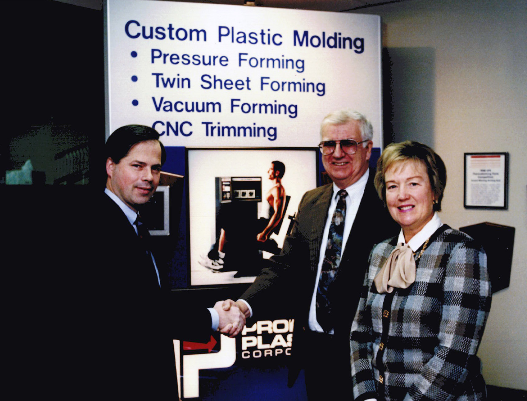 The Grundys on the right and Steve Murrill on the left, Steve and John are shaking hands. In the background is a sign that reads "Custom Plastic Molding * Pressure Forming * Twin Sheet Forming * Vacuum Forming * CNC Trimming"