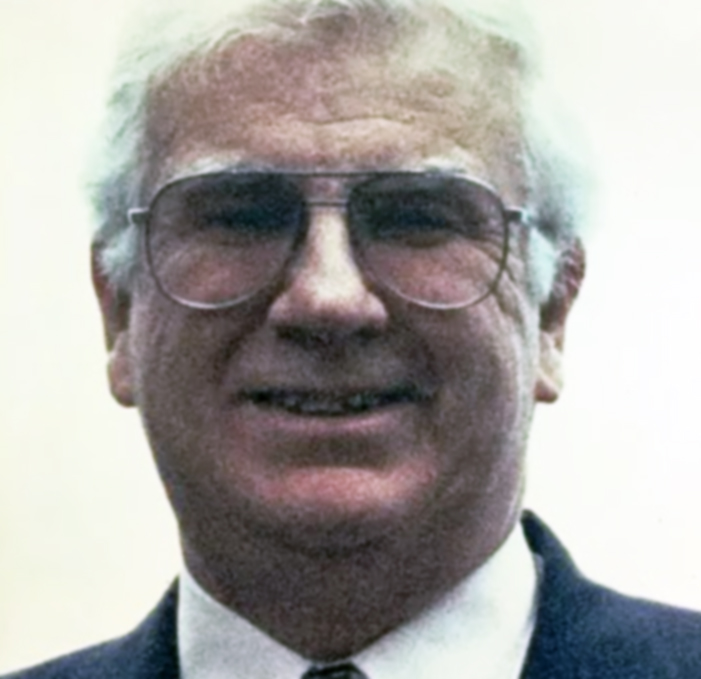 John Grundy headshot circa 1993, John has white hair and glasses, and is wearing a blue suit and tie.