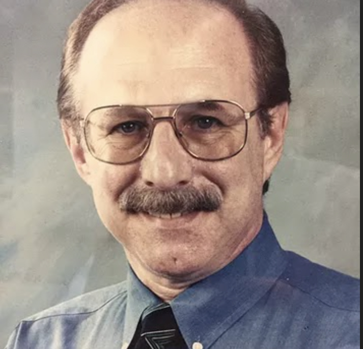 Stephen Sweig headshot, Stephen has brown thinning hair, a mustache, and large rimmed glasses. He's wearing a blue shirt and dark striped tie in front of a professional photography background.