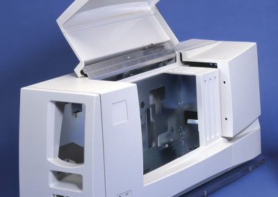 White ID card printer machine with an open top lid and exposed interior, shown on a blue background. The front has a card slot and ventilation openings.